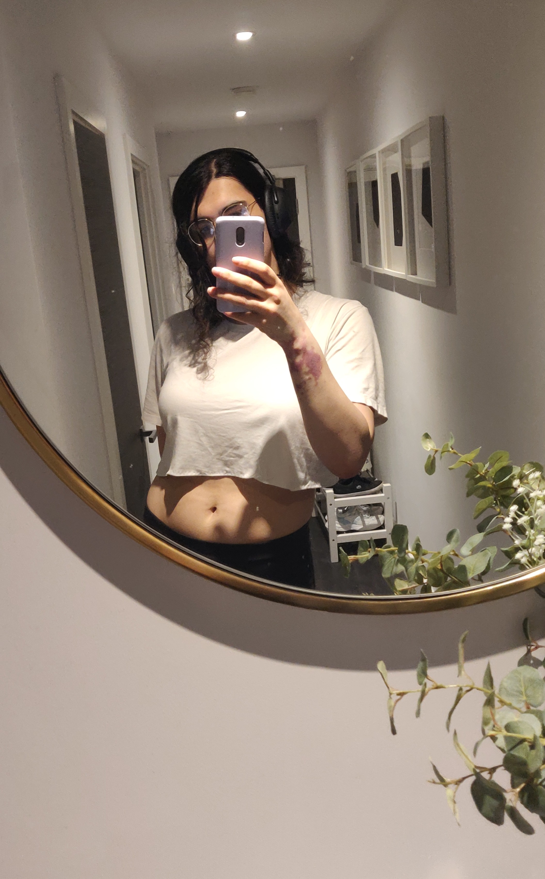 annieversary selfie on a mirror at the end of a corridor. shes wearing a white crop top, black pants, glasses that dont actually glass, and black headphones because she has autism. her hair is tied up with a hairclaw, so you cant see most of it 