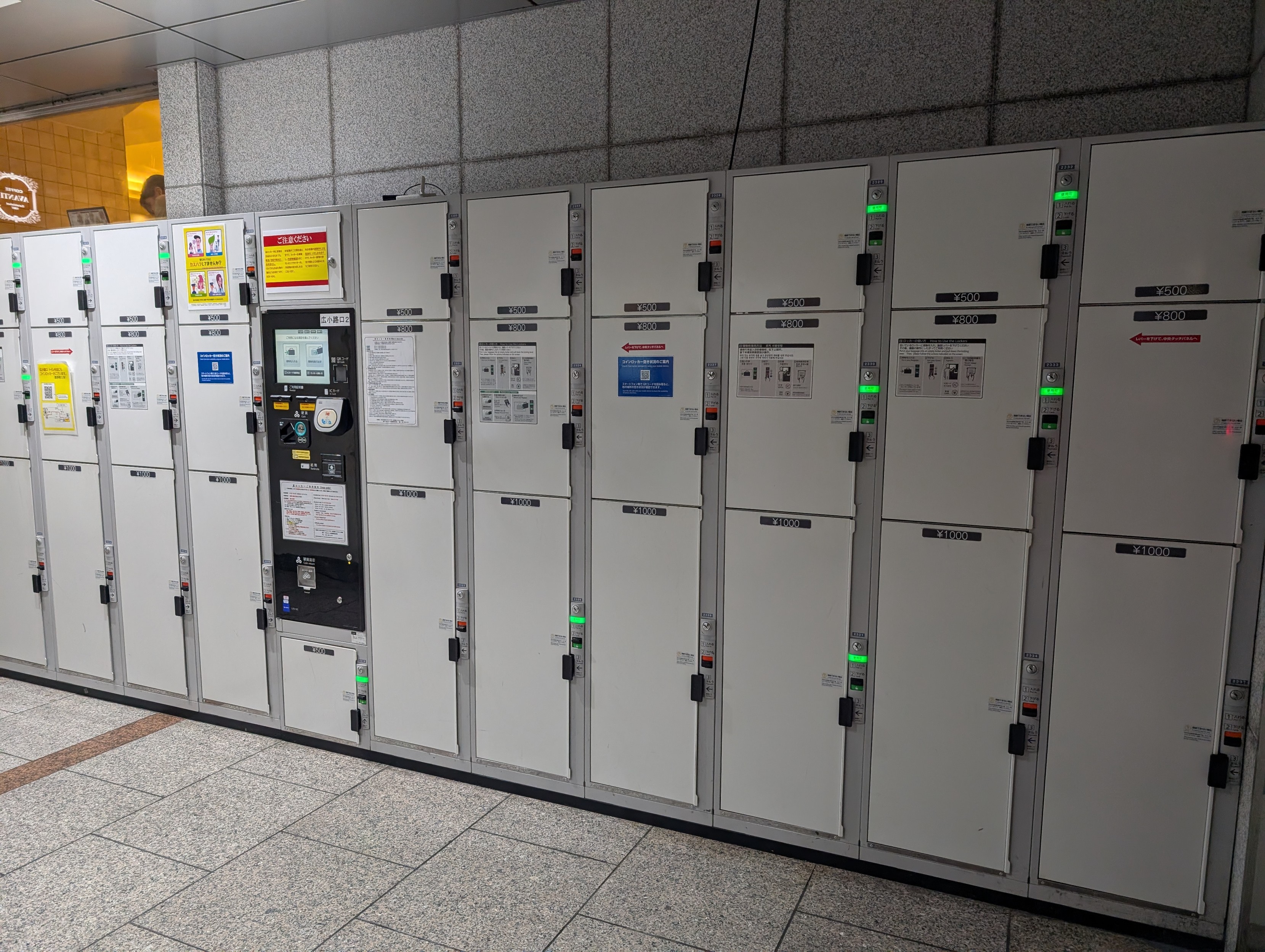 A line up of metal luggage lockers of varying sizes. Some have green LEDs to indicate their availability 