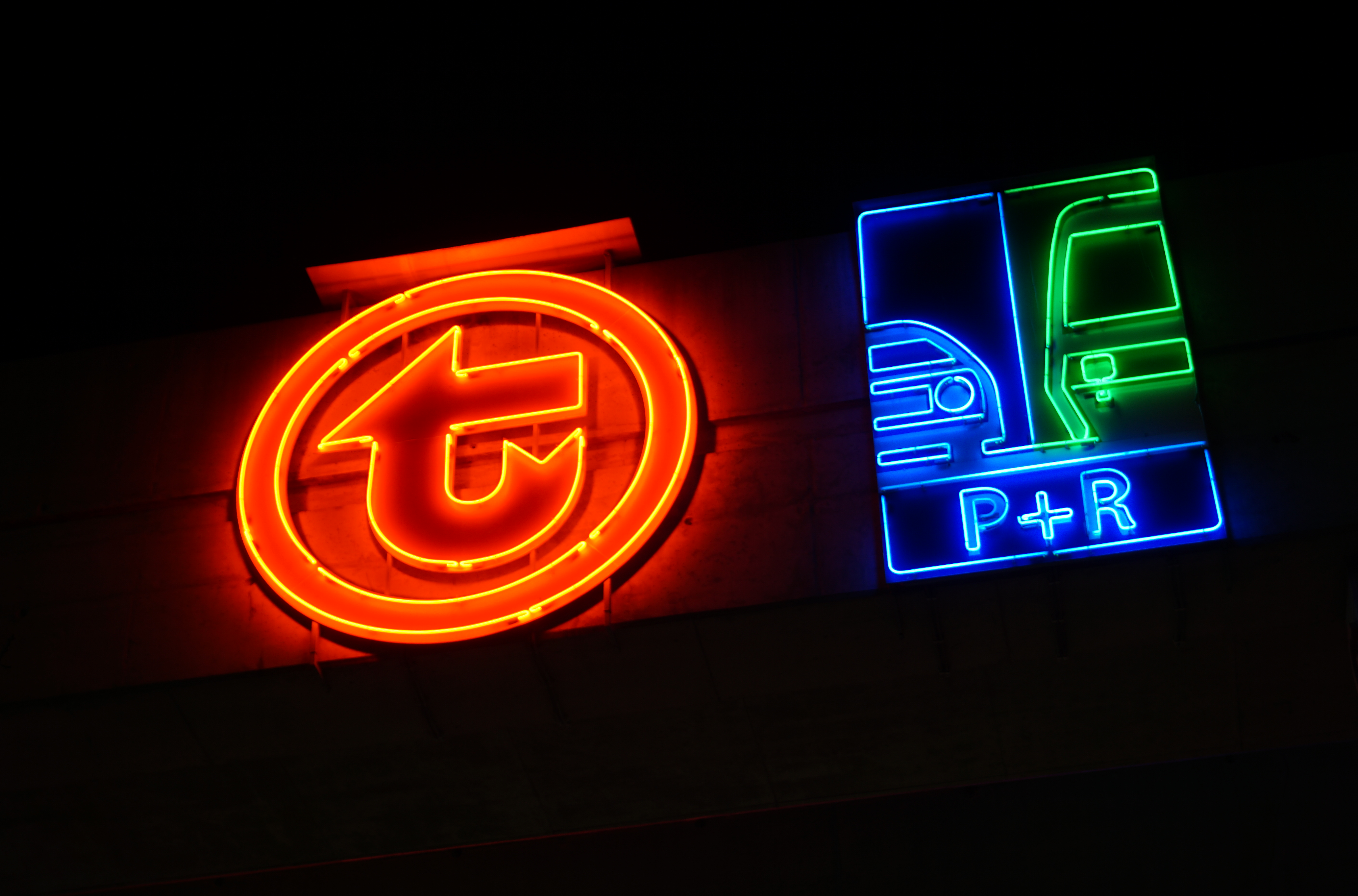 red neon with Warsaw Public Transport logo next to the blue and green P+R logo, both on the wall, nothing besides them visible