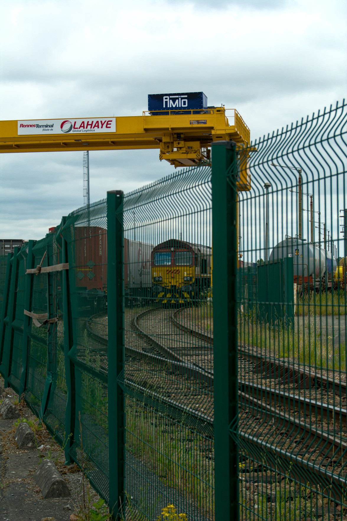 Behind a green mesh fence, until a container loading system, a british class 66 locomotive is parked in a rail yard. Behind a green mesh fence, until a container loading system, a british class 66 locomotive is parked in a rail yard.