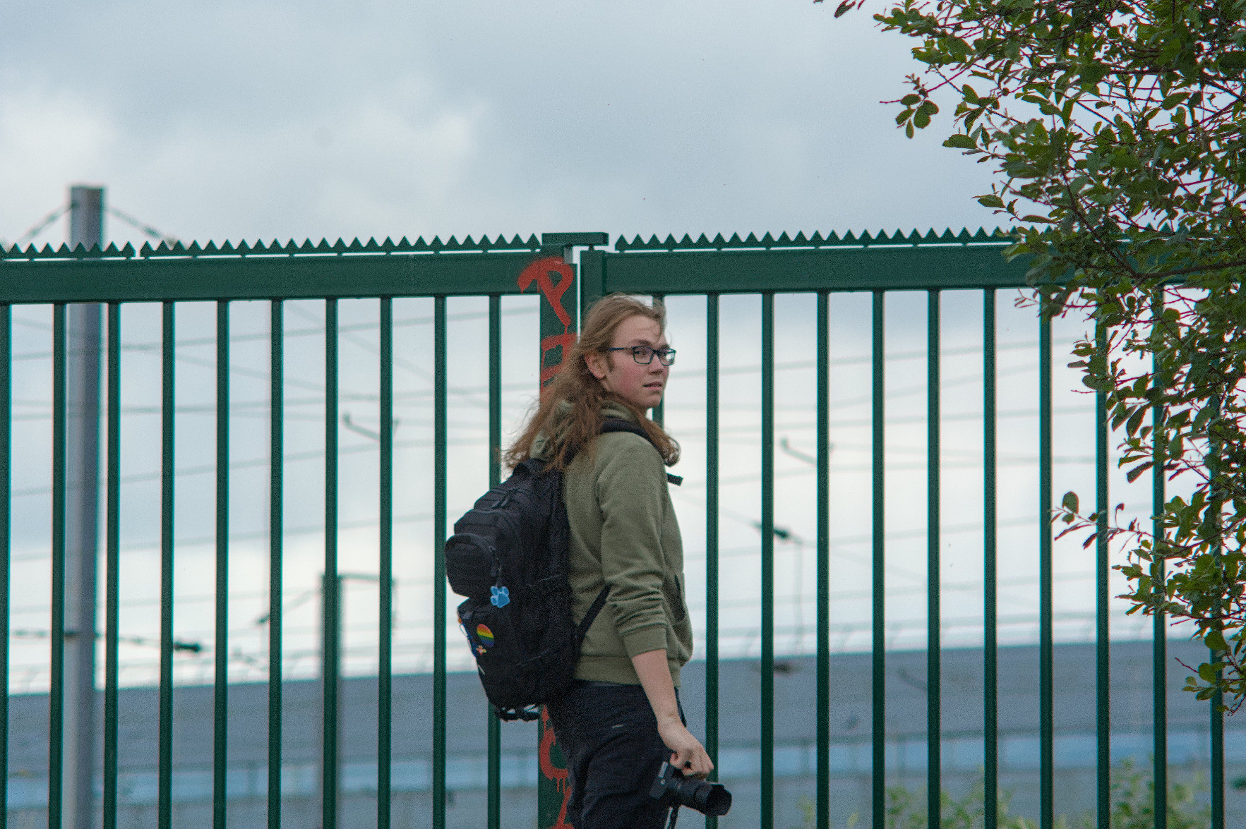 @rail@flufftech.net looking back to the photographer. They’re holding their camera and facing a green fence overlooking the rail yard. @rail@flufftech.net looking back to the photographer. They’re holding their camera and facing a green fence overlooking the rail yard.