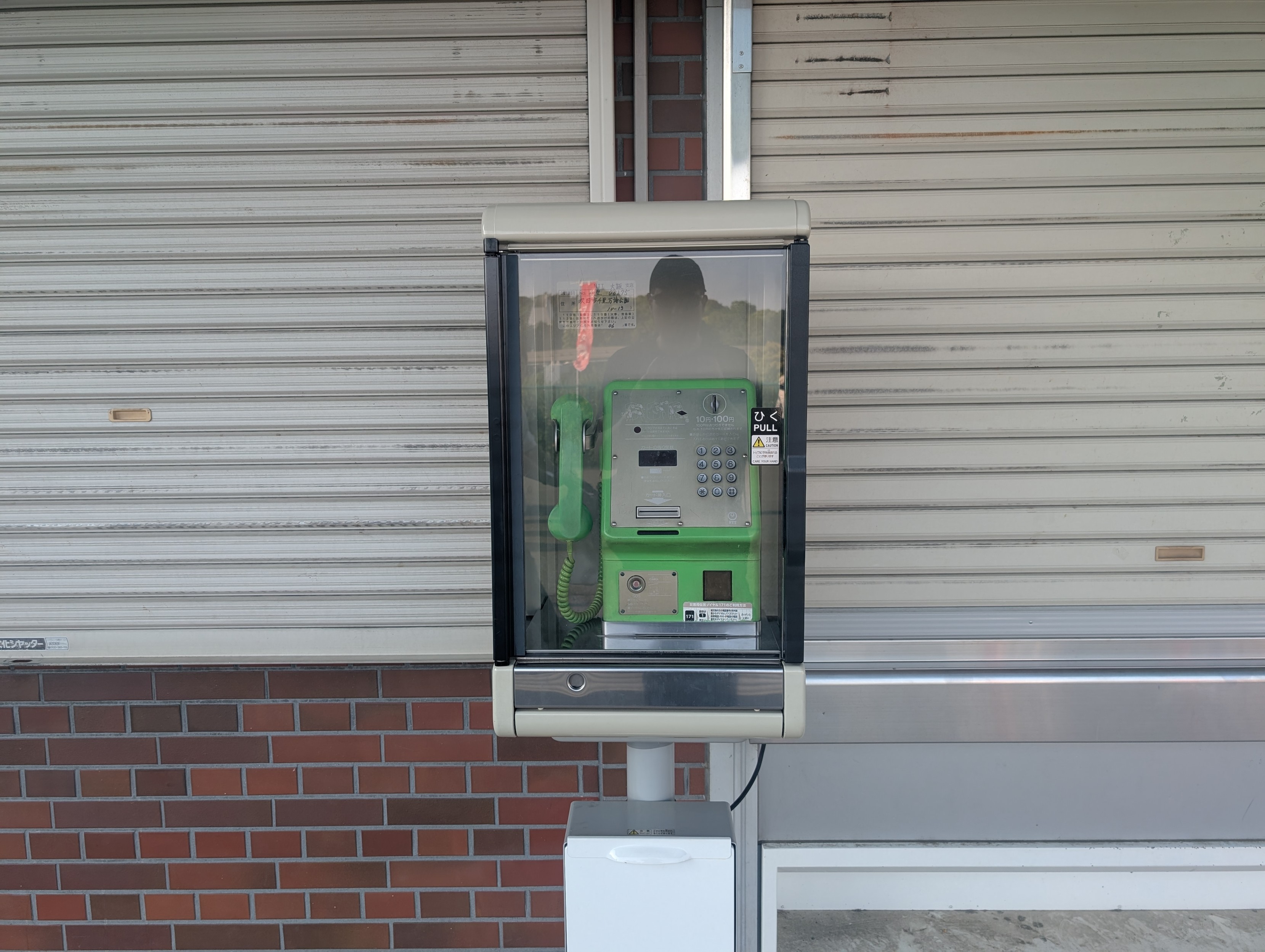 A green NTT telephone in a sort of box, with some closed shutters behind it. 