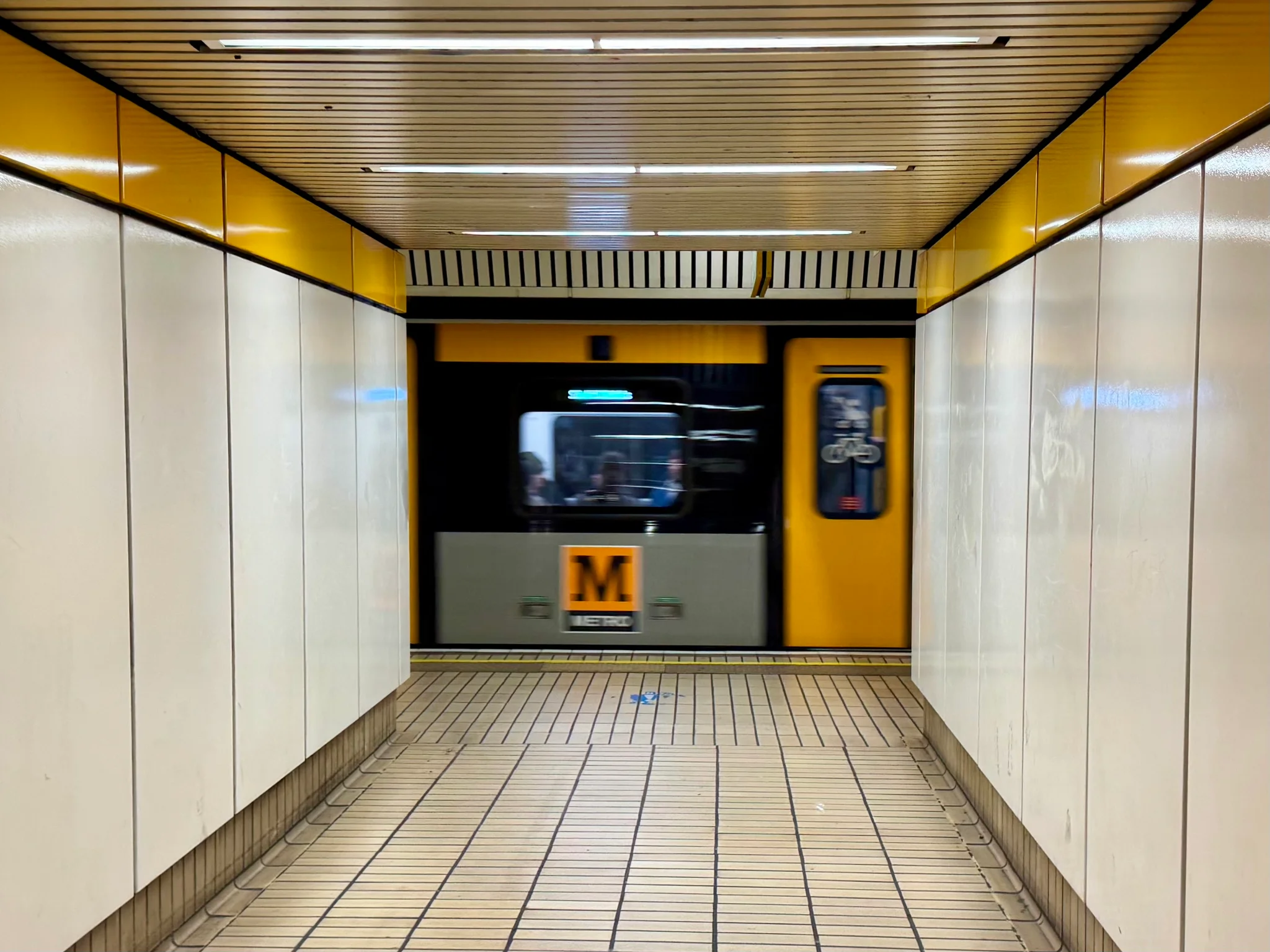 A metro vehicle departing from the track opposite, framed by a connection tunnel with white panelling and yellow accents.