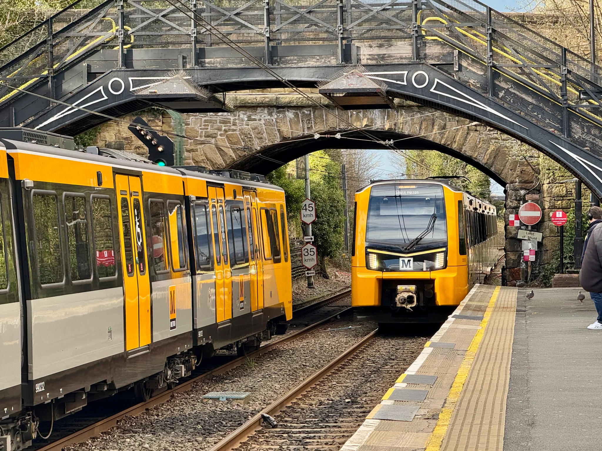 A modern Class 555 Stadler Metro in mostly orangy-yellow vehicle approaching South Gosforth station, with another vehicle standing on the far track. There are two bridges over the railway here, one iron footbridge, and one stone bridge.