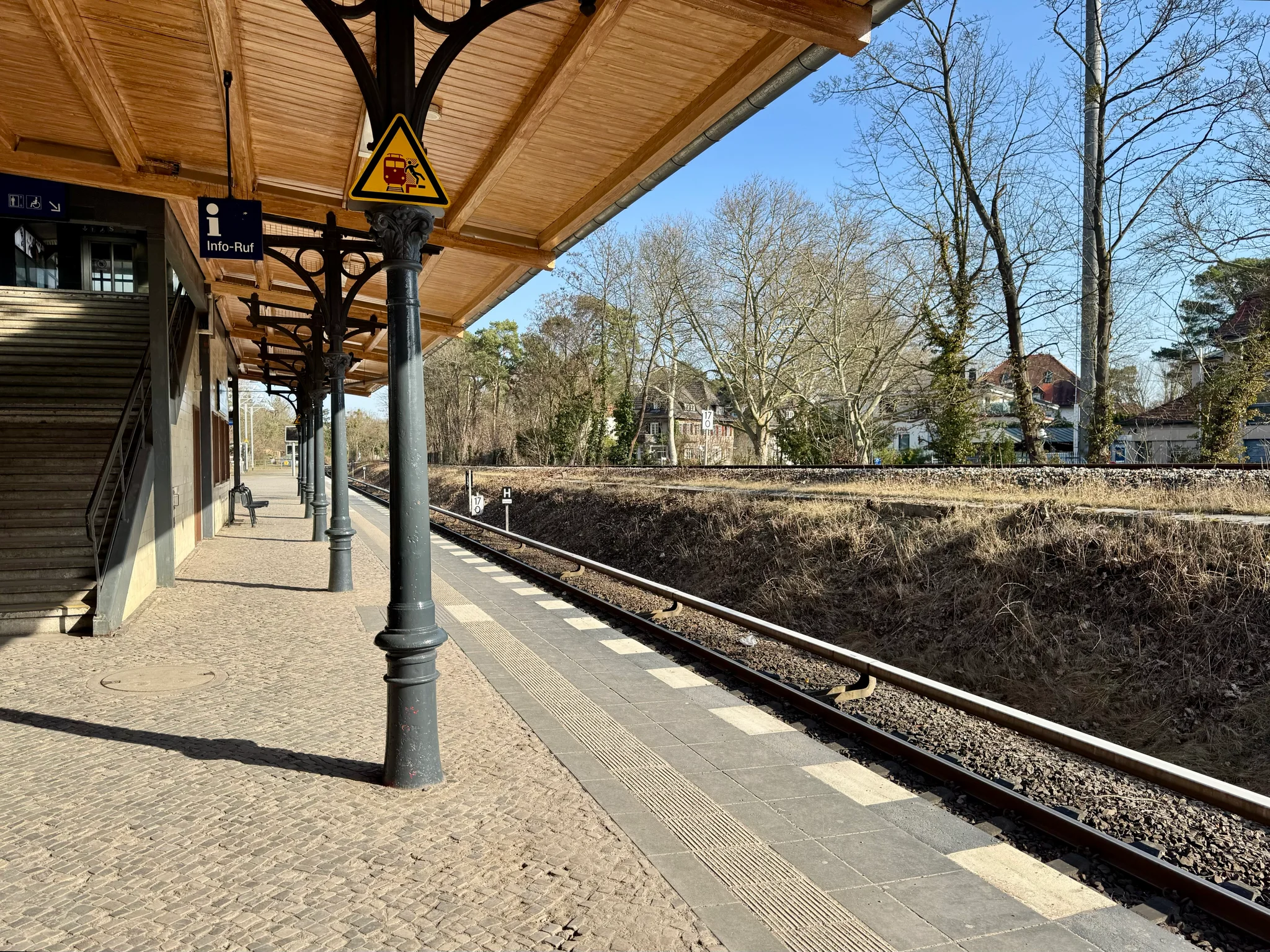 Station Nikolassee’s platform with a wooden canopy and an earth wall behind the S-Bahn tracks with more infrastructure on it.