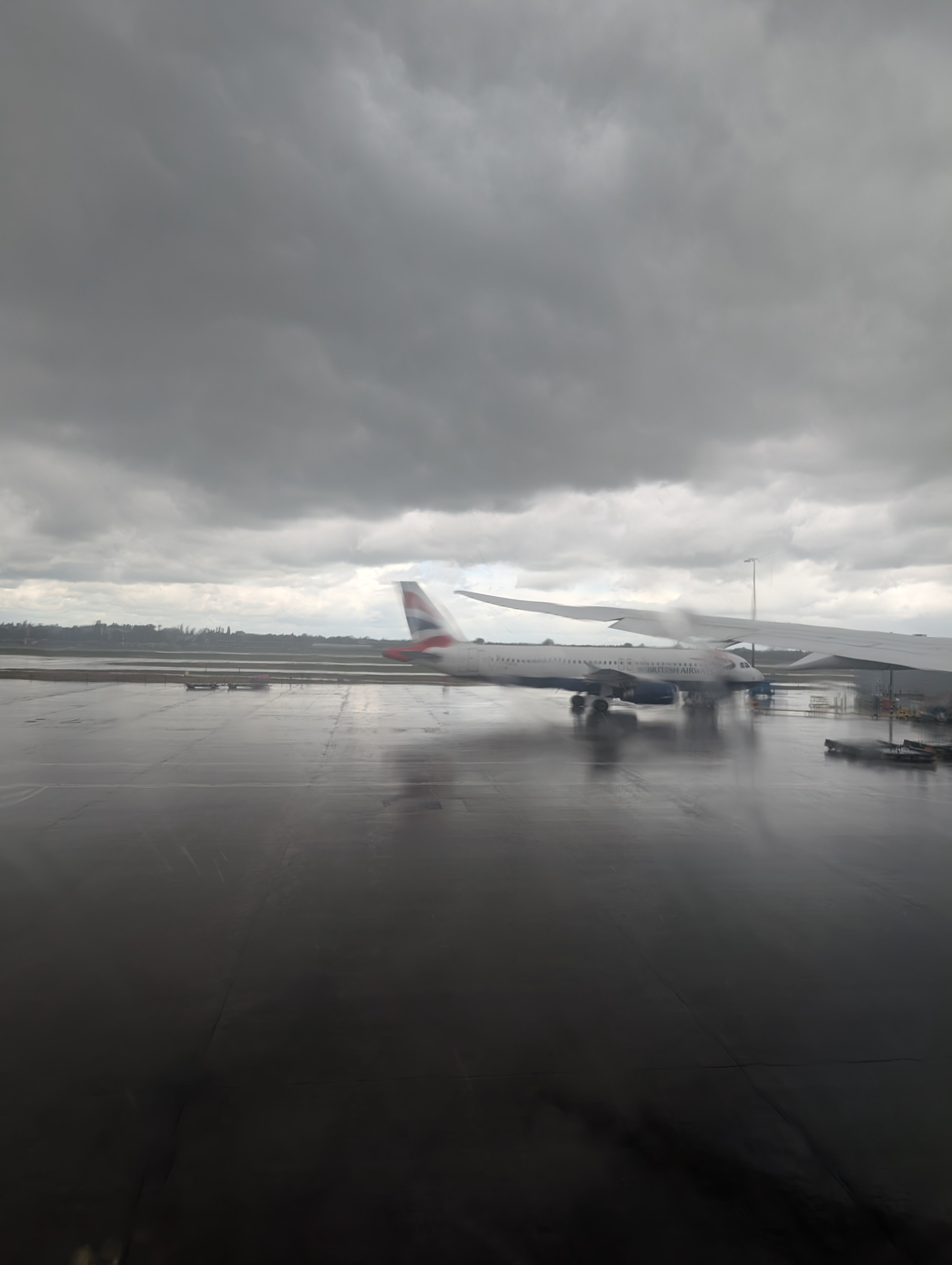 An A320 sits on the tarmac with brooding skies above and water pooling on the ground. The photos is taken from an aircraft, and distorted by the rain 