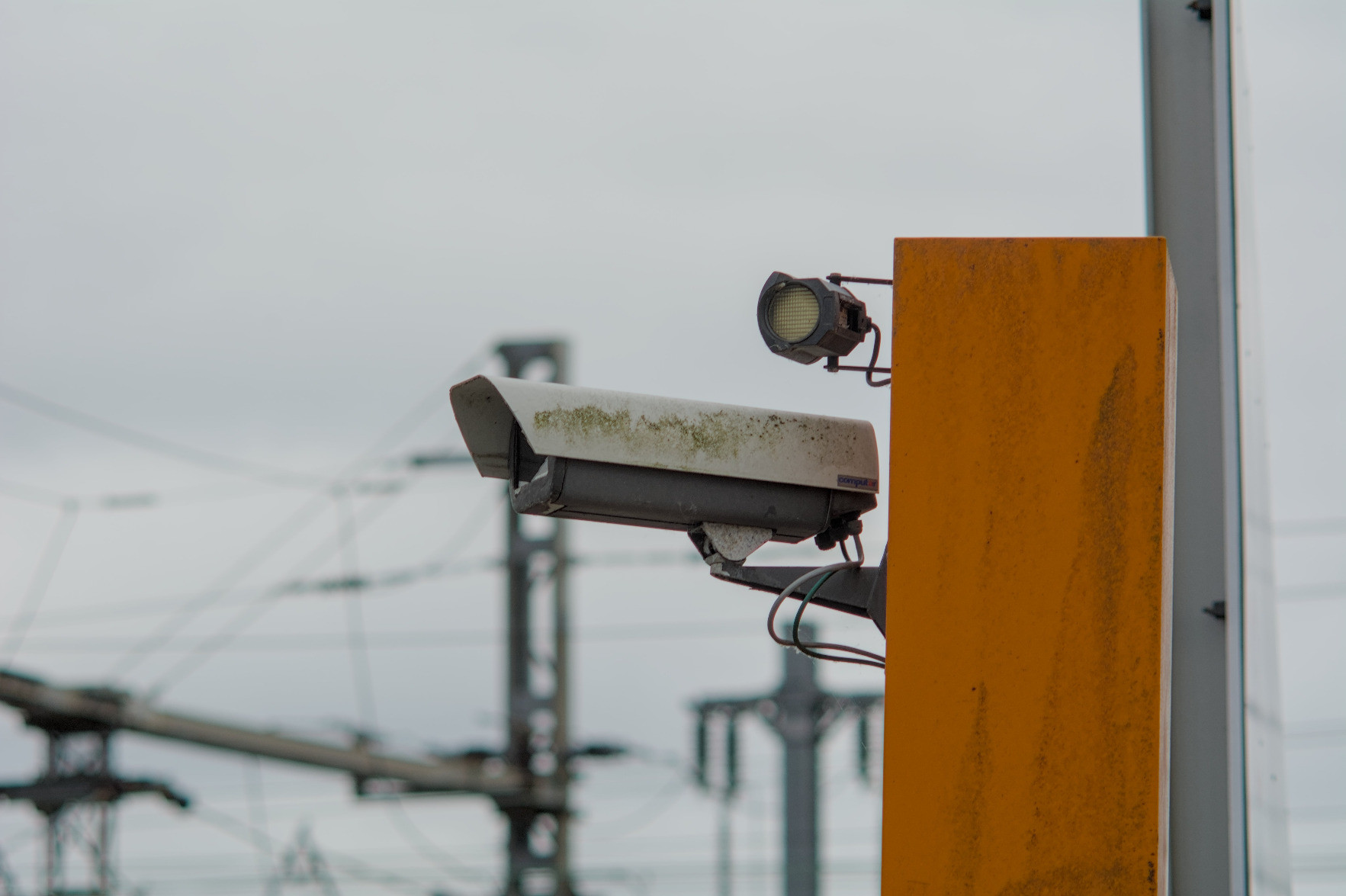 A security camera at the entrance of the rail yard; blurry catenaries and poles are visible in the background. A security camera at the entrance of the rail yard; blurry catenaries and poles are visible in the background.