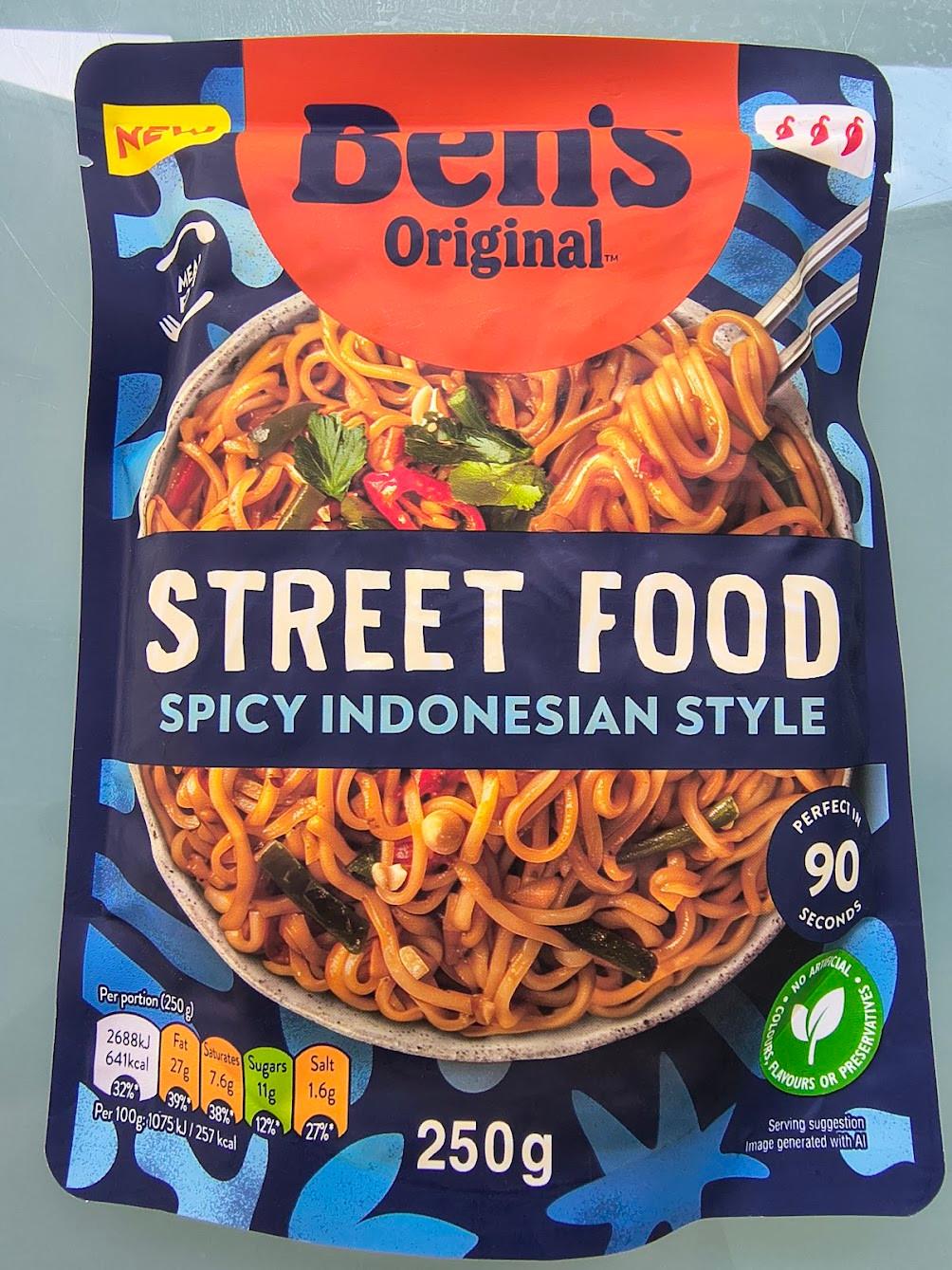 A photograph of a package of Ben's Original Street Food Spicy Indonesian Style noodles, sold in the UK. The package is dominated by a picture of a bowl of noodles, with vegetable-looking things in them. A tiny, tiny note to the bottom-right reads "Serving suggestion. Image generated with AI." A photograph of a package of Ben's Original Street Food Spicy Indonesian Style noodles, sold in the UK. The package is dominated by a picture of a bowl of noodles, with vegetable-looking things in them. A tiny, tiny note to the bottom-right reads "Serving suggestion. Image generated with AI."