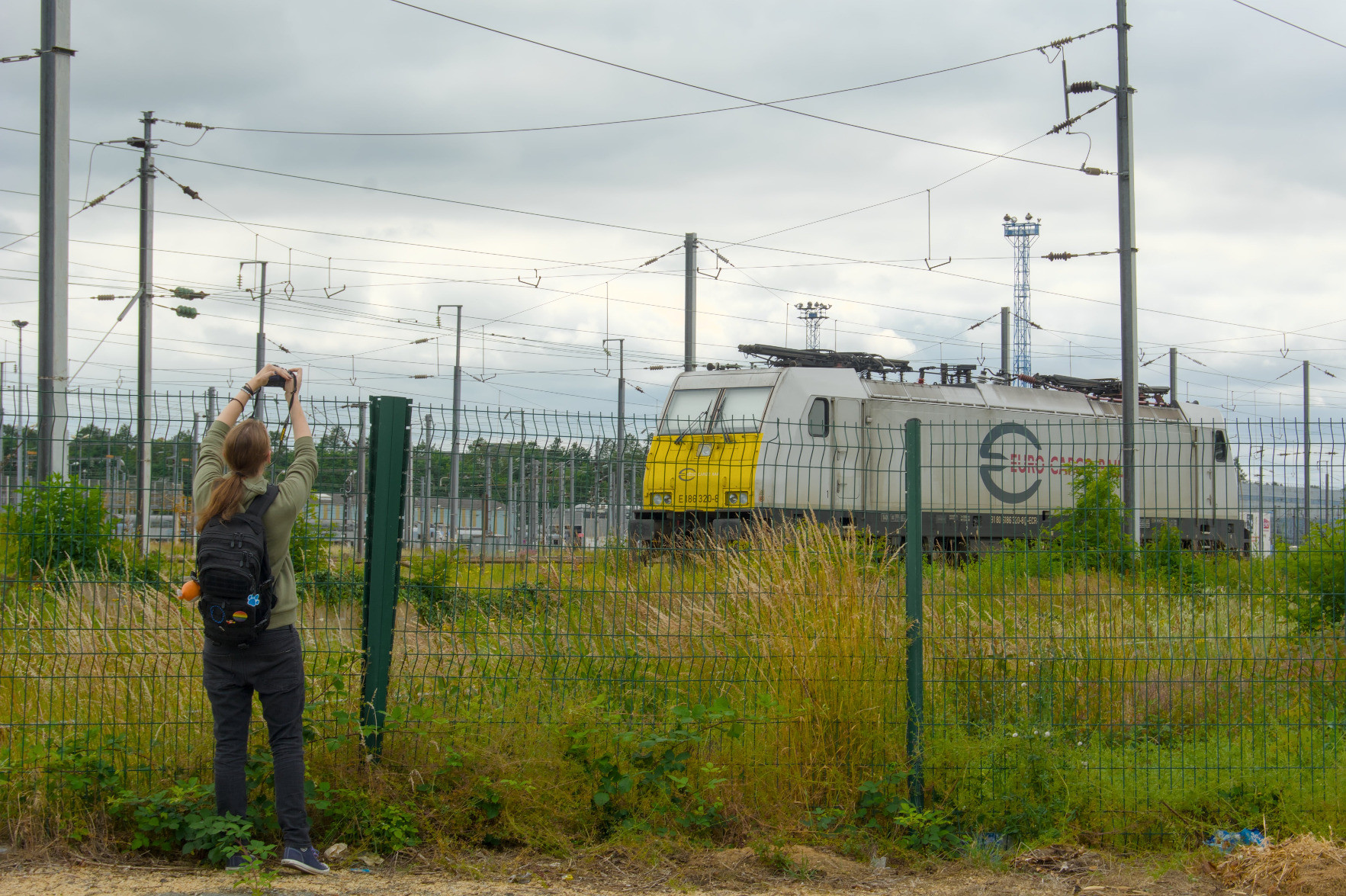 @rail@flufftech.net in front of a fence with their camera pointed above it, they’re taking a photo of the cargo train behind the fence. @rail@flufftech.net in front of a fence with their camera pointed above it, they’re taking a photo of the cargo train behind the fence.