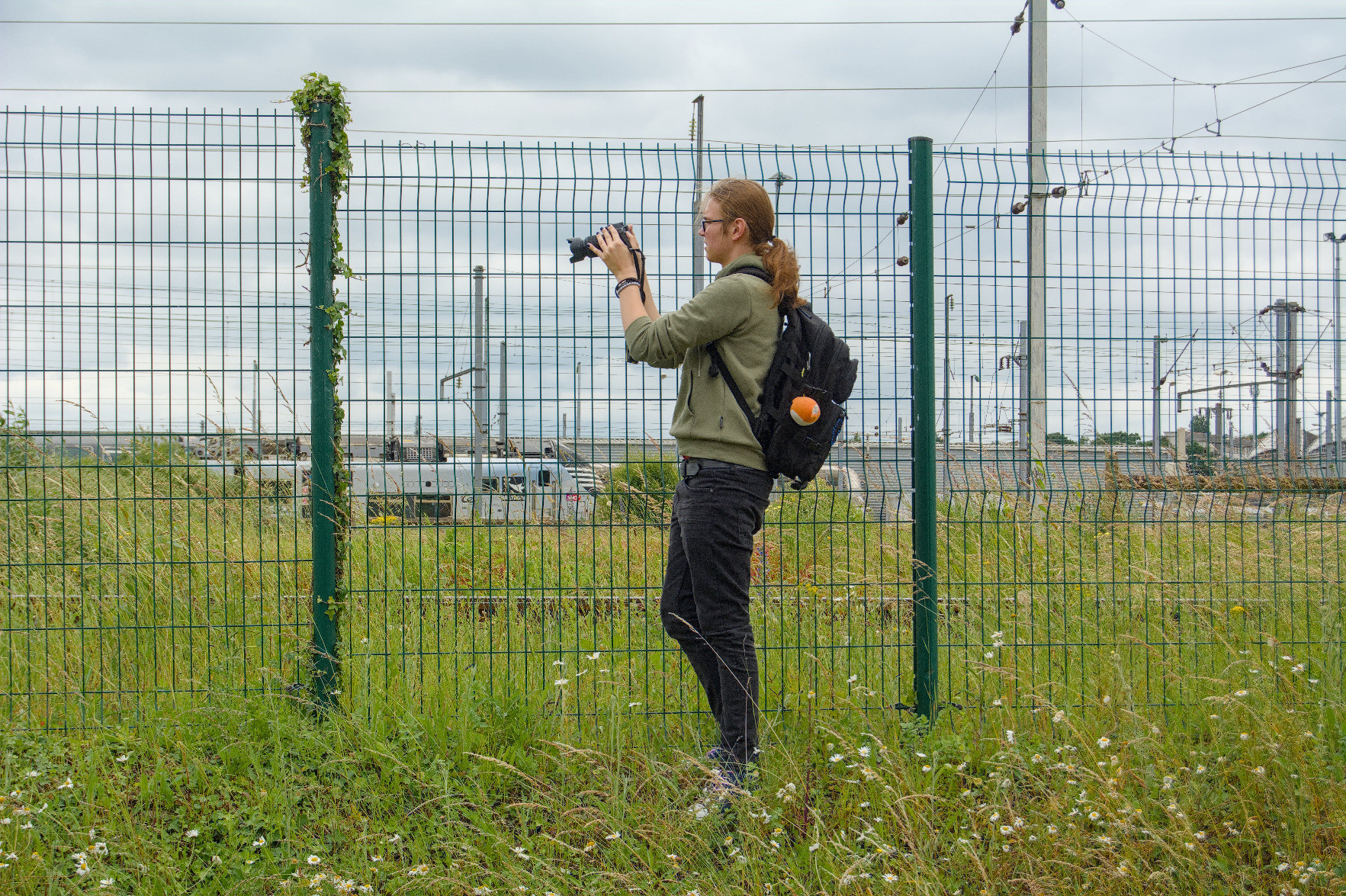 @rail@flufftech.net taking a photo with their camera; trains are visible in the background behind a green mesh fence. @rail@flufftech.net taking a photo with their camera; trains are visible in the background behind a green mesh fence.