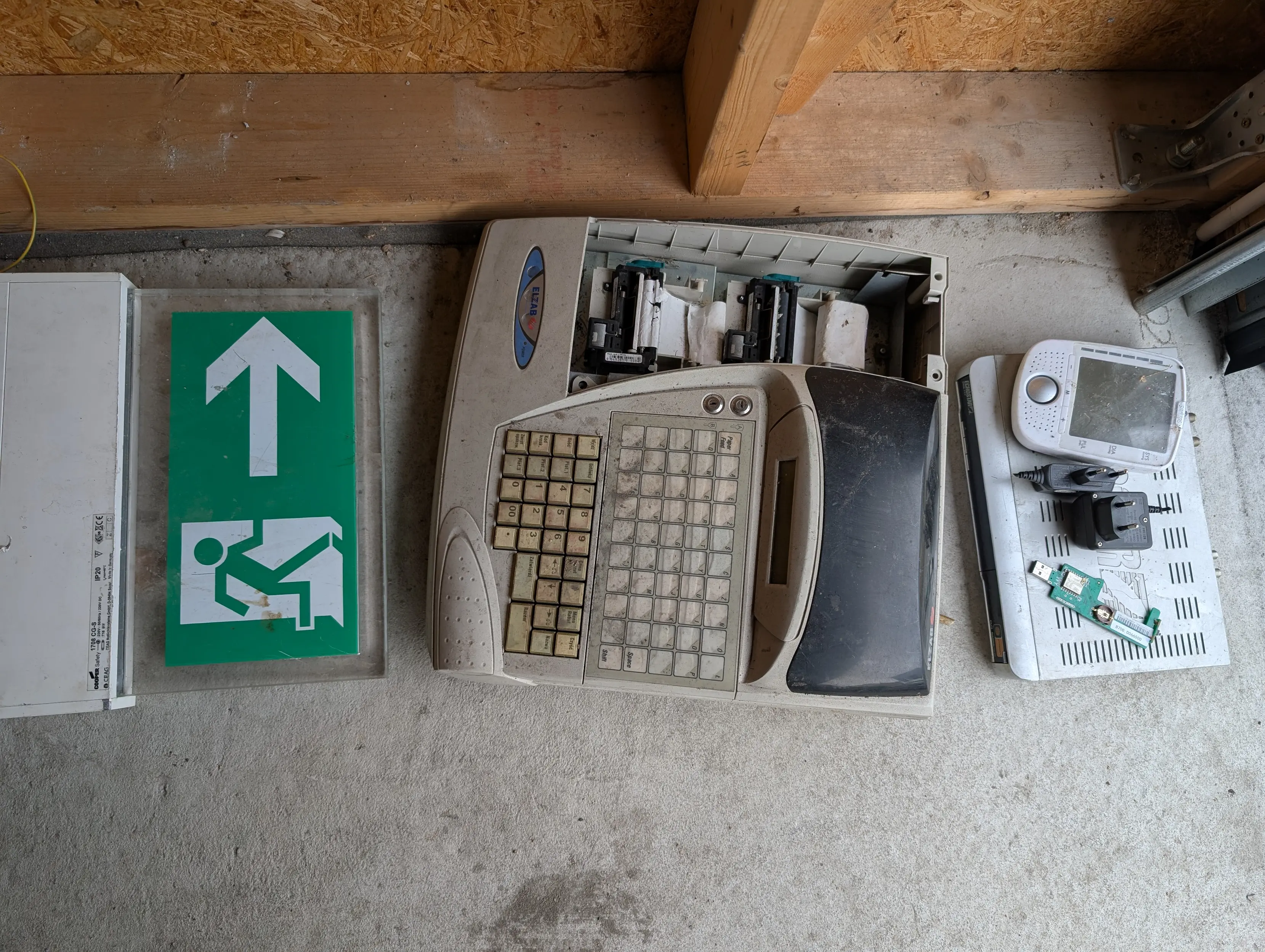 from right to left: a satellite set top box, some usb dongle with an esp8266 module, two cable-less chargers, only one of which i intended to take, a blood pressure meter, and then a literal elzab brand cash register complete with a mechanical keypad and Two thermal printer assemblies, And an actual emergency exit sign the kind that gets hung above doors and stuff and glows