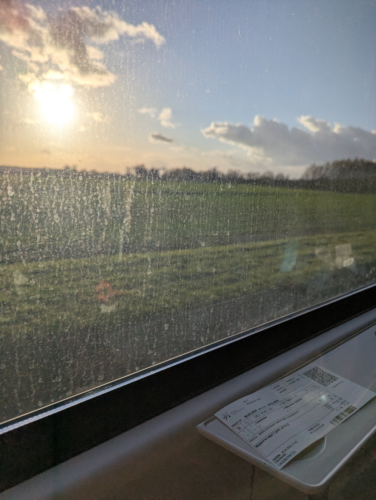 View out of a dirty train window during sunset in rural Mazovia. In front of the window there's a paper ticket for that train