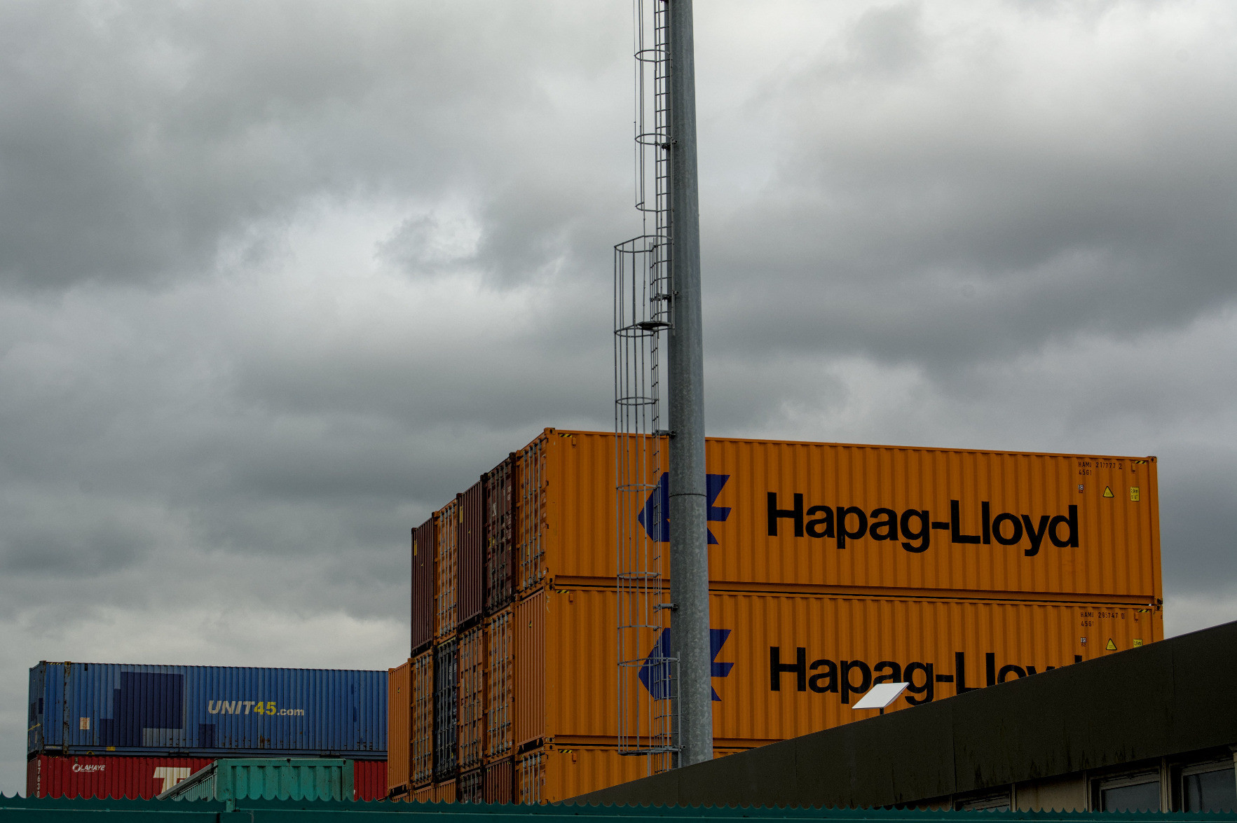 Containers piled up behind a building and a big pole. The sky, cloudy and grey, contrasts with the colourful Hapag-Lloyd containers. Containers piled up behind a building and a big pole. The sky, cloudy and grey, contrasts with the colourful Hapag-Lloyd containers.