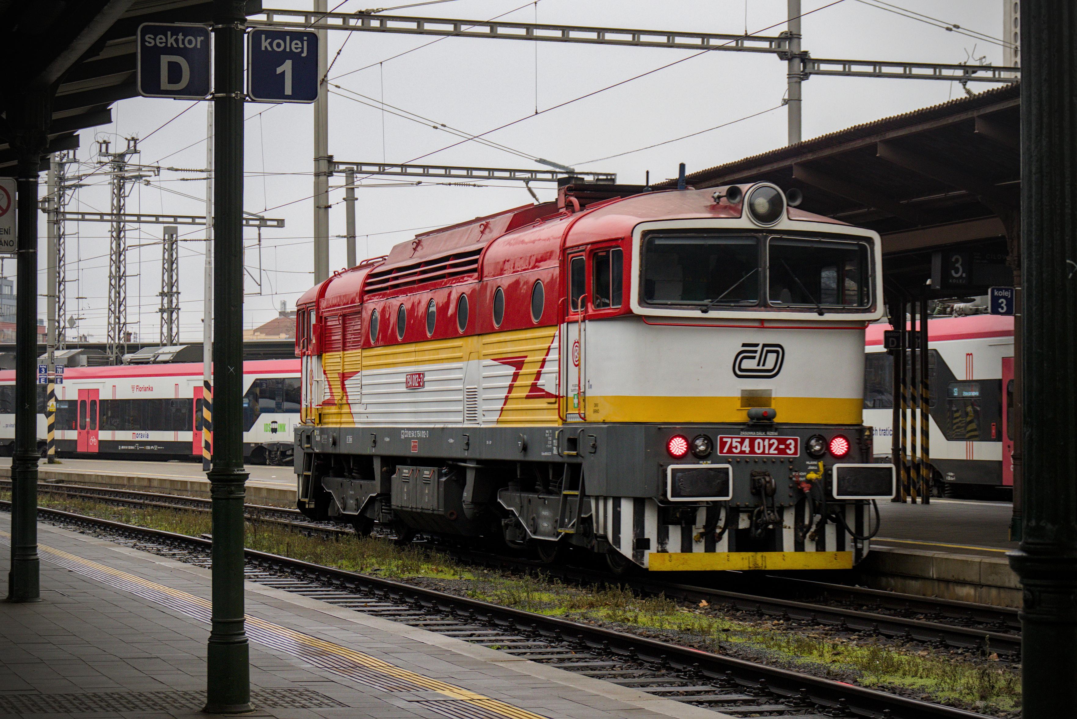 a ČD class 754 locomotive in a red-yellow-white livery with some zigzag shaped decals on the side, departing from Brno hl.n.